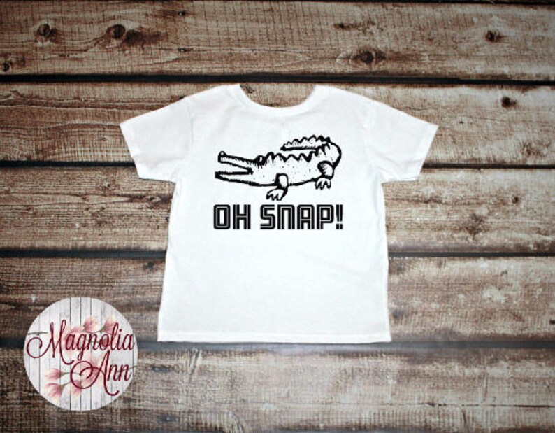 May include: White t-shirt with a black and white illustration of an alligator and the text "Oh Snap!"