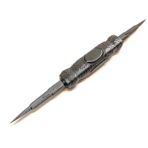 May include: A black, intricately designed tool with a pointed tip on each end. The tool has a decorative, textured handle with a circular detail in the center.