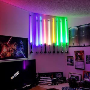 May include: A wall display of 12 colorful lightsabers mounted on a wall with a black and white Star Wars poster and a TV screen in the background.