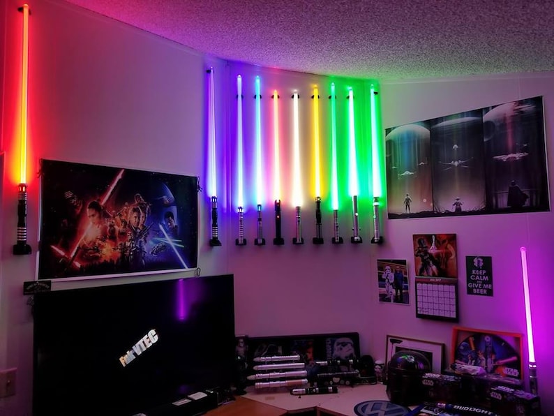 May include: A wall display of 12 colorful lightsabers mounted on a wall with a black and white Star Wars poster and a TV screen in the background.