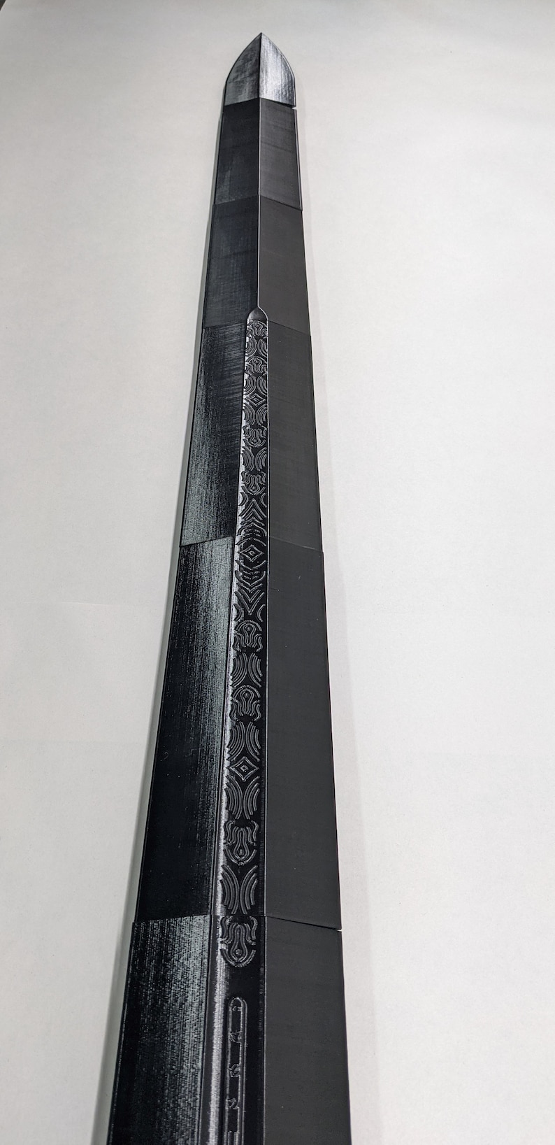K&ouml;nnte beinhalten: Ein schwarzes Schwert mit einem detaillierten Design auf der Klinge. Das Schwert ist aus Metall und hat eine scharfe Spitze.