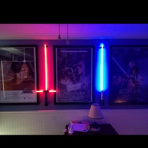 May include: Two light-up lightsaber props mounted on a wall, one red and one blue. The lightsabers are in front of three movie posters, "Star Wars", "The Empire Strikes Back", and "Return of the Jedi".