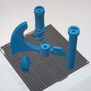 May include: A collection of blue 3D-printed components, possibly for a model or toy, arranged on a black grid. The parts include a handle, a blade-like shape, and other geometric forms, suggesting a construction project.
