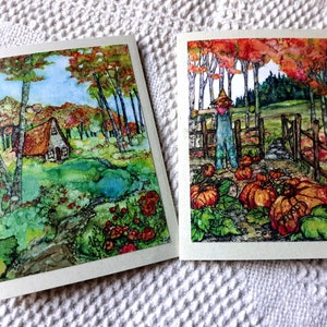 May include: Two watercolor paintings on cards. The first card shows a small cottage in a forest with a stream running through it. The second card shows a scarecrow standing in a pumpkin patch.