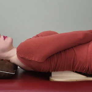 May include: A woman lying on her back on a red surface with a brown pillow under her head. She is wearing a red shirt and black pants. A wooden wedge is under her back.