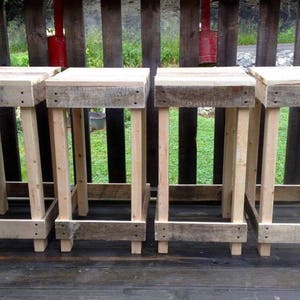 May include: Four rustic wooden stools with a natural finish. Each stool has a square seat and four sturdy legs, connected by a lower frame. The stools are made from reclaimed wood, showcasing the wood's natural grain and texture.