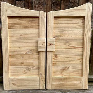 May include: Two wooden saloon-style doors with a natural finish. Each door has a rectangular frame with a curved top and horizontal planks. The doors are joined by a wooden latch. The wood grain is visible.