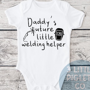 May include: White baby onesie with black text that reads "Daddy's future little welding helper". The onesie has a black illustration of a welding mask and a welding torch.
