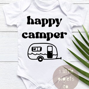 May include: White baby onesie with black text that reads "happy camper" and a black and white illustration of a camper. The text "3 little piglets co." is in the bottom right corner.