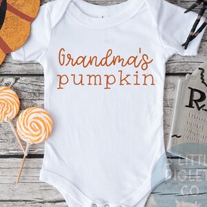 K&ouml;nnte beinhalten: Wei&szlig;er Baby-Strampler mit dem Text "Grandma's pumpkin" in orangefarbener Schrift. Der Strampler liegt auf einem Holzgrund mit einem K&uuml;rbis, S&uuml;&szlig;igkeiten und einem Grabstein.