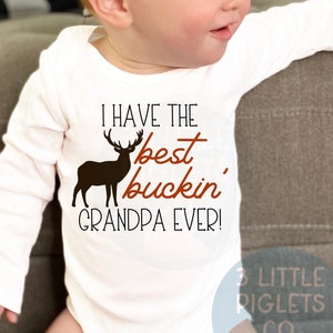 May include: White long-sleeve onesie with a brown deer silhouette and the text "I have the best buckin' grandpa ever!" in orange. The onesie has a small logo in the bottom right corner that says "3 Little Piglets Co."