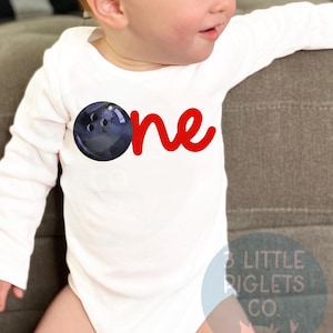 May include: White long-sleeve onesie with a red "one" and a blue bowling ball graphic. The onesie is worn by a baby.