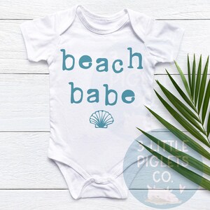 May include: White baby bodysuit with teal text reading 'beach babe' and a teal seashell graphic.  The bodysuit is shown on a white wooden surface with a green tropical leaf.