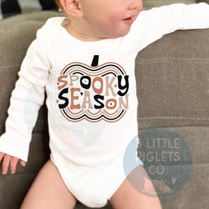 Puede incluir: Un body blanco de manga larga para beb&eacute; con un gr&aacute;fico de calabaza marr&oacute;n, negro y blanco que dice "Spooky Season".