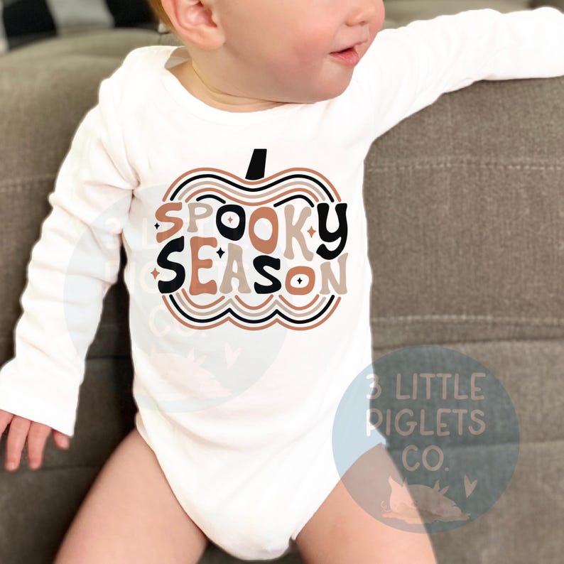 Puede incluir: Un body blanco de manga larga para beb&eacute; con un gr&aacute;fico de calabaza marr&oacute;n, negro y blanco que dice "Spooky Season".