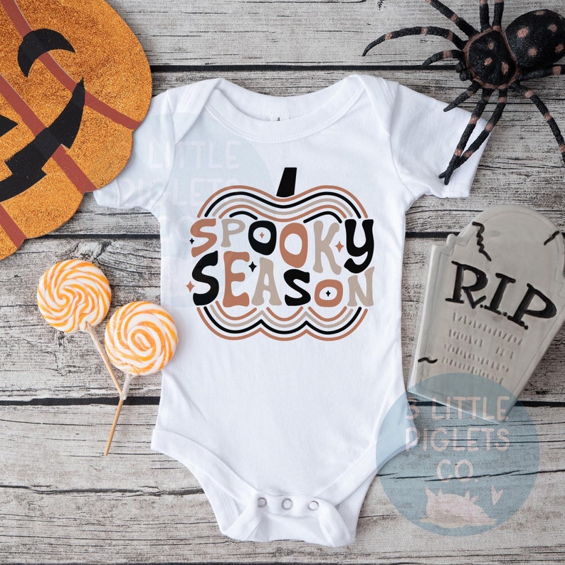Puede incluir: Un body blanco para beb&eacute; con un dise&ntilde;o de calabaza marr&oacute;n, naranja y negro. La calabaza tiene el texto "Spooky Season" en su interior.