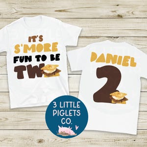 May include: Two white short-sleeve t-shirts. The left shirt features the text "IT'S S'MORE FUN TO BE TWO" in brown and black with a s'more graphic. The right shirt displays the name "DANIEL" in yellow, a large brown number "2", and a s'more graphic.