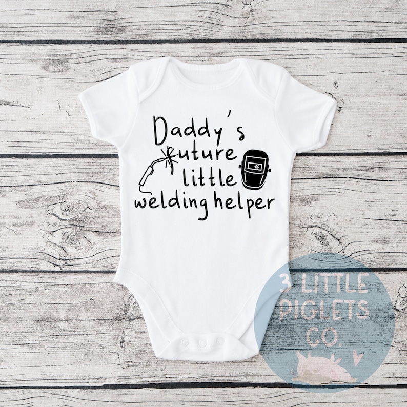 May include: White baby onesie with black text that reads "Daddy's future little welding helper". The onesie has a black illustration of a welding mask and a welding torch.
