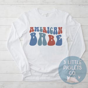 May include: White long-sleeve shirt with the text "AMERICAN BABE" in blue, red, and teal retro font.