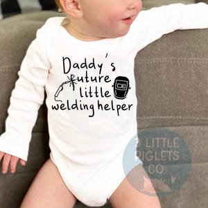May include: White long-sleeve baby onesie with black text that reads "Daddy's future little welding helper" and a black illustration of a welding mask and welding torch.