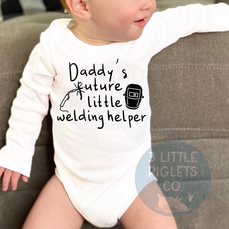 May include: White long-sleeve baby onesie with black text that reads "Daddy's future little welding helper" and a black illustration of a welding mask and welding torch.