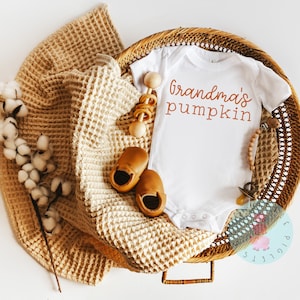 K&ouml;nnte beinhalten: Ein wei&szlig;er Baby-Strampler mit dem Text "Grandma's pumpkin" in orangefarbener Schrift. Der Strampler liegt in einem Weidenkorb mit einer braunen Decke, einer Holzrassel und einem Paar braunen Babyschuhen.