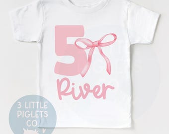 May include: White t-shirt with a pink number 5 and a pink bow. The name "River" is written below the number.