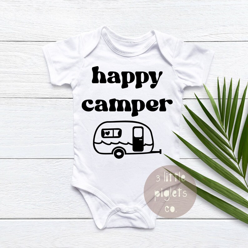 May include: White baby onesie with black text that reads "happy camper" and a black and white illustration of a camper. The text "3 little piglets co." is in the bottom right corner.