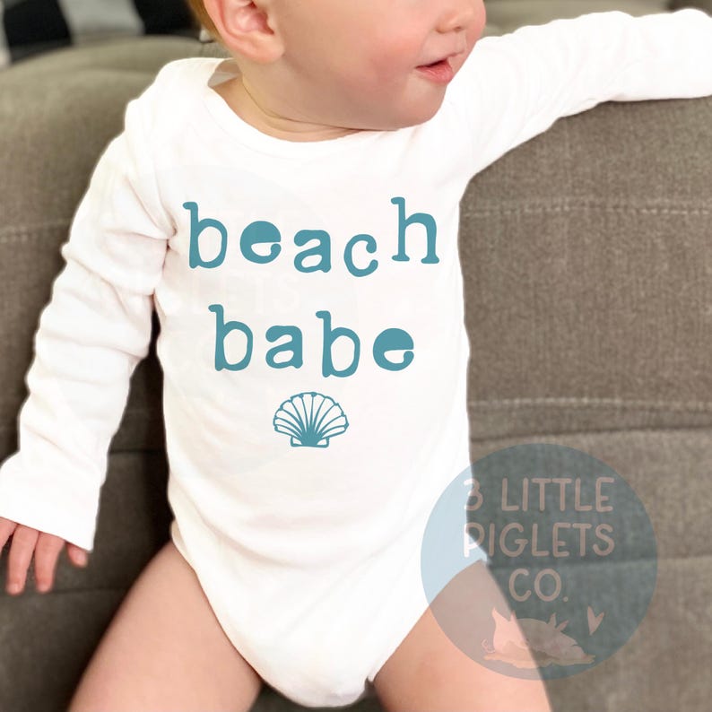 May include: A white baby bodysuit with long sleeves, featuring the teal text "beach babe" and a seashell graphic. The garment is made from a soft fabric. The brand logo is visible in the bottom right corner of the bodysuit.