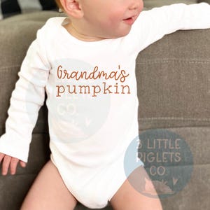 K&ouml;nnte beinhalten: Wei&szlig;er Langarm-Strampler mit dem Text "Grandma's pumpkin" in Orange. Der Strampler wird von einem Baby getragen.