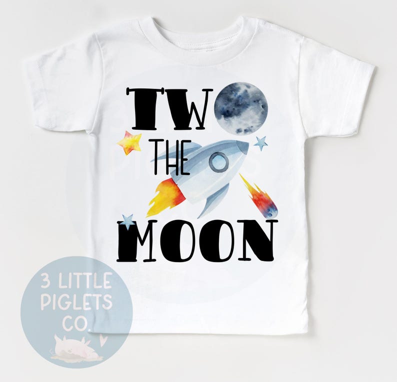 May include: White t-shirt with a colorful graphic featuring a rocket ship, moon, and stars. The text "TWO THE MOON" is displayed in black. A small logo for "3 LITTLE PIGLETS CO." is at the bottom.