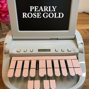 May include: A white Stenograph machine with a black screen displaying the text "PEARLY ROSE GOLD". The machine has a row of pink keys with a textured surface.