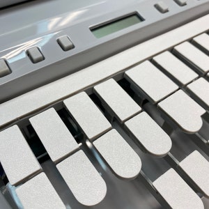 May include: Close-up of a light gray portable typewriter with white keys. The keys are arranged in a curved row, with a small display screen and several buttons above the keys. The typewriter has a textured surface.