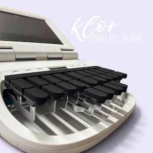 May include: A white stenograph machine featuring a row of black rectangular keys with metallic supports. The device has a screen and the text Klor STENO KEY DESIGN is visible in the upper right corner of the frame.