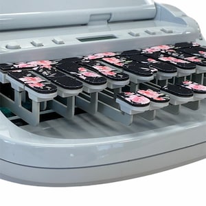 May include: A white electric typewriter with a floral patterned keyboard. The keys are black with pink flowers.