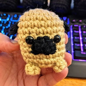 May include: A crocheted pug dog toy with black eyes and a black nose. The toy is beige and has a textured surface.