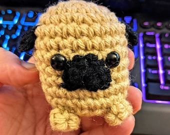 May include: A crocheted pug dog toy with black eyes and a black nose. The toy is beige and has a textured surface.