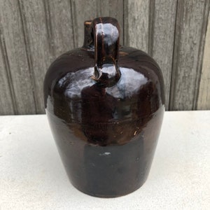 May include: A dark brown glazed stoneware jug with a handle. The jug has a rounded body and a slightly flared lip.