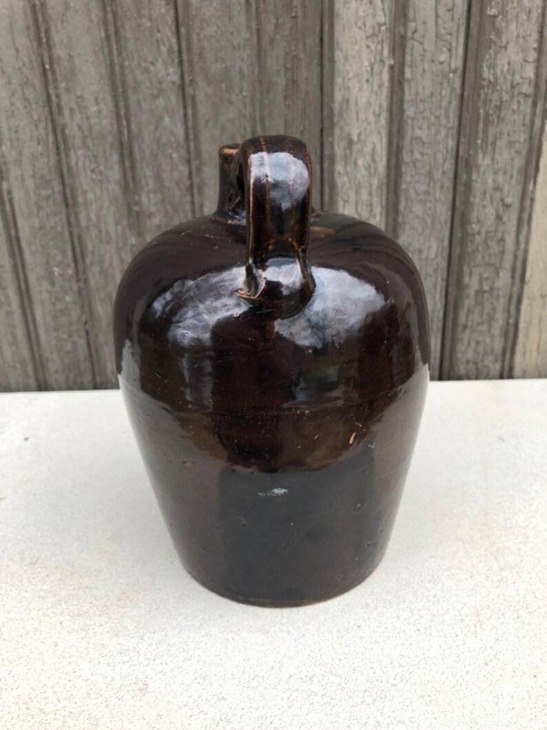 May include: A dark brown glazed stoneware jug with a handle. The jug has a rounded body and a slightly flared lip.