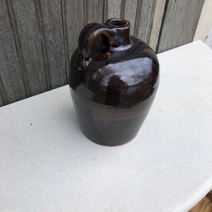 May include: A dark brown glazed stoneware jug with a single handle. The jug has a rounded body and a small opening at the top.