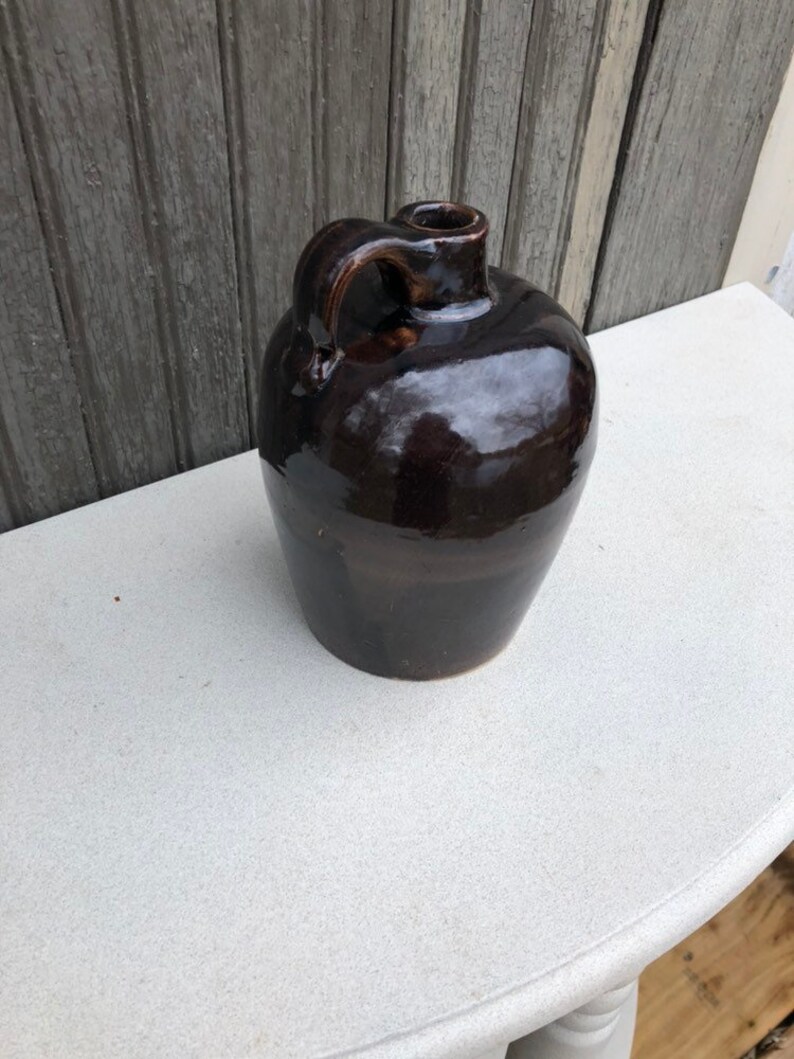 May include: A dark brown glazed stoneware jug with a single handle. The jug has a rounded body and a small opening at the top.