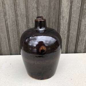 May include: A dark brown ceramic bottle with a rounded body and a narrow neck. The bottle has a glossy finish and a slight sheen.