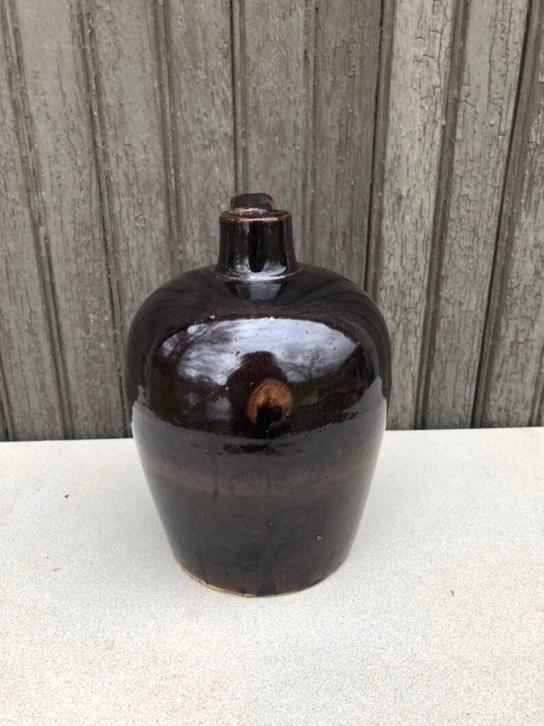 May include: A dark brown ceramic bottle with a rounded body and a narrow neck. The bottle has a glossy finish and a slight sheen.