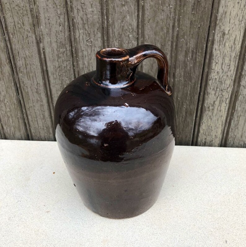 May include: A dark brown glazed stoneware jug with a rounded body and a single handle. The jug has a smooth, glossy finish.