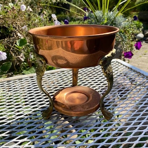 May include: A copper fondue pot with a brass stand and three legs. The pot has a round shape and a smooth surface. The stand has a decorative design with a floral motif.