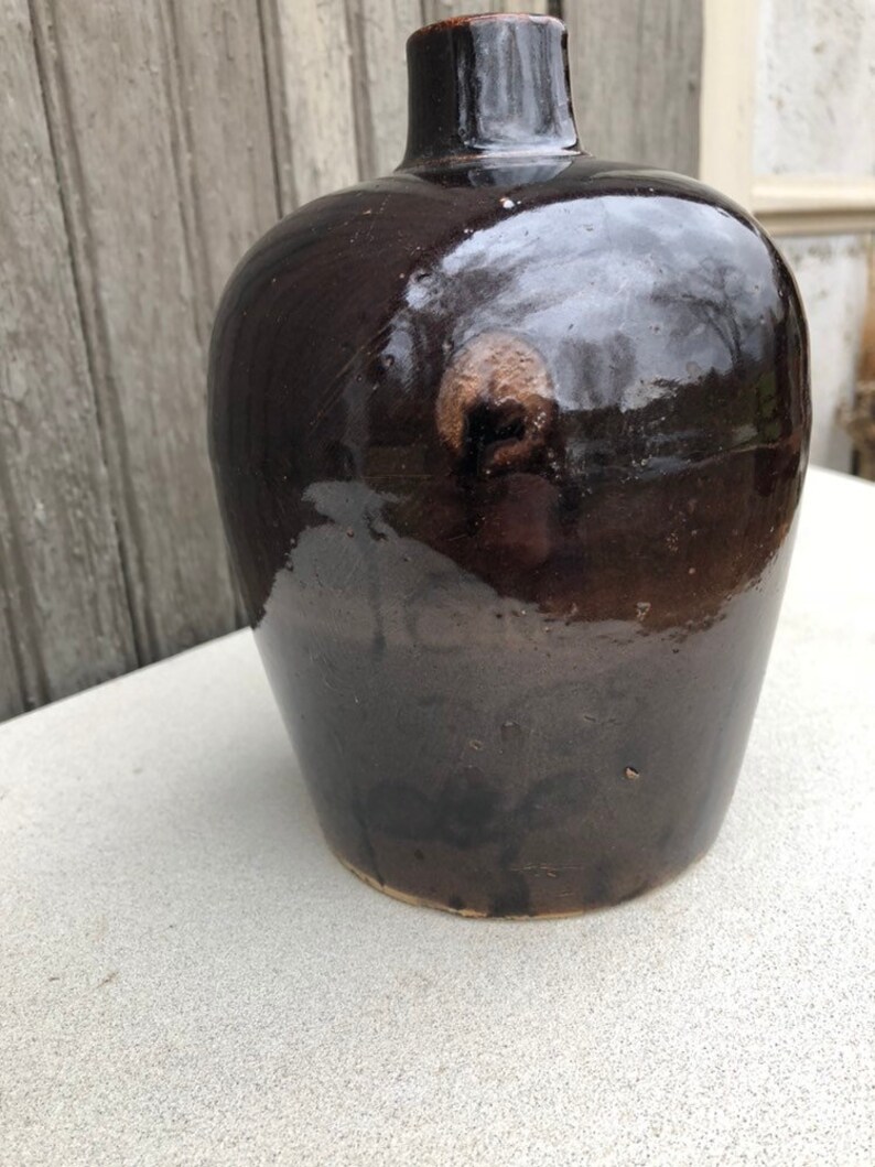 May include: A dark brown ceramic vase with a rounded body and a narrow neck. The vase has a glossy finish and a subtle pattern of brown and black.