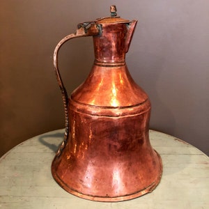 May include: A large, antique copper pitcher with a wide, bell-shaped body and a curved handle. The pitcher has a hammered finish and a slightly tarnished patina.