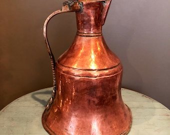 May include: A large, antique copper pitcher with a wide, bell-shaped body and a curved handle. The pitcher has a hammered finish and a slightly tarnished patina.