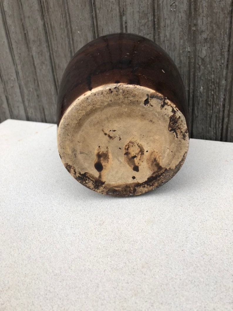 May include: A brown glazed ceramic crock with a light brown base. The crock has a rounded shape and a wide opening.
