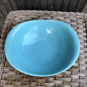 May include: A large, oval-shaped, light blue ceramic bowl with a speckled glaze. The bowl is sitting on a woven wicker surface.